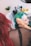 A person is applying hair dye to another person's hair in a salon setting. The person receiving the dye has green hair, while the one applying it has red hair. Various hair products and tools are visible in the background.