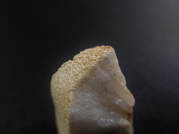 A close-up of a rough, beige mineral or stone with a textured surface and jagged edges against a dark, blurred background.