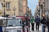 A busy street scene with several people walking along a narrow road lined with buildings. In the foreground, there is a small three-wheeled vehicle with a 'APE TAXI' sign, carrying a few passengers. The background features a row of buildings on both sides of the road, street lamps, and mountains in the far distance.