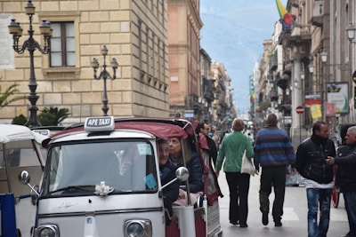 A busy street scene with several people walking along a narrow road lined with buildings. In the foreground, there is a small three-wheeled vehicle with a 'APE TAXI' sign, carrying a few passengers. The background features a row of buildings on both sides of the road, street lamps, and mountains in the far distance.