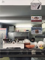 A rustic kitchen or diner environment features hanging signs, including those for Coca-Cola, pizza, and hamburgers. There is a vintage weighing scale on the counter, with a basket of snacks and some stacked plastic containers in view. A person is partially visible in the background near industrial kitchen equipment.