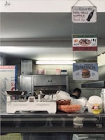 A rustic kitchen or diner environment features hanging signs, including those for Coca-Cola, pizza, and hamburgers. There is a vintage weighing scale on the counter, with a basket of snacks and some stacked plastic containers in view. A person is partially visible in the background near industrial kitchen equipment.