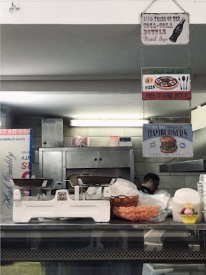 A rustic kitchen or diner environment features hanging signs, including those for Coca-Cola, pizza, and hamburgers. There is a vintage weighing scale on the counter, with a basket of snacks and some stacked plastic containers in view. A person is partially visible in the background near industrial kitchen equipment.