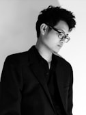 A person with short, dark hair is wearing glasses and a dark suit jacket over a dark shirt. The lighting is soft, creating a shadow on the white background, giving the image a contemplative feel.