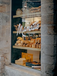 A bakery display window featuring an assortment of freshly baked bread and pastries. Several varieties of bread are arranged on wooden shelves inside the store. The glass window is adorned with the logo and name of the bakery, and there are wicker baskets and a crate visible in the display.