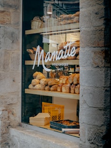 A bakery display window featuring an assortment of freshly baked bread and pastries. Several varieties of bread are arranged on wooden shelves inside the store. The glass window is adorned with the logo and name of the bakery, and there are wicker baskets and a crate visible in the display.