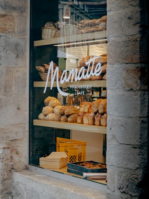 A bakery display window featuring an assortment of freshly baked bread and pastries. Several varieties of bread are arranged on wooden shelves inside the store. The glass window is adorned with the logo and name of the bakery, and there are wicker baskets and a crate visible in the display.