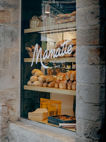 A bakery display window featuring an assortment of freshly baked bread and pastries. Several varieties of bread are arranged on wooden shelves inside the store. The glass window is adorned with the logo and name of the bakery, and there are wicker baskets and a crate visible in the display.
