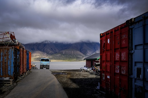 A rugged landscape with overcast skies dominating the background. In the foreground, there are large shipping containers on both sides of a narrow path. A large blue truck is parked near the edge of a tranquil body of water, with mountainous terrain in the distance.