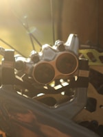 A close-up of a metallic bicycle disc brake system, highlighted by sunlight. The intricate details of the brake caliper and rotor are visible, showcasing the precision engineering. The background is softly blurred, enhancing the focus on the mechanical elements.