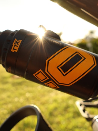 A close-up shot of a black water bottle with a large yellow 'O' and additional yellow designs. The bottle is secured to a bicycle frame, with sunlight shining and lens flare visible near the top. The background features bright green grass and blurred outdoor scenery.