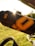 A close-up shot of a black water bottle with a large yellow 'O' and additional yellow designs. The bottle is secured to a bicycle frame, with sunlight shining and lens flare visible near the top. The background features bright green grass and blurred outdoor scenery.