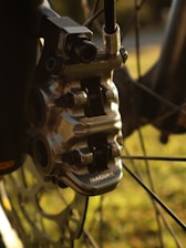 A close-up view of a metallic bicycle brake caliper, showcasing intricate engineering details with the brand name 'Magura' visible. The background appears soft and blurred, hinting at a natural outdoor setting with hints of green and yellow coloring.