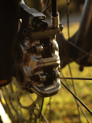 A close-up view of a metallic bicycle brake caliper, showcasing intricate engineering details with the brand name 'Magura' visible. The background appears soft and blurred, hinting at a natural outdoor setting with hints of green and yellow coloring.
