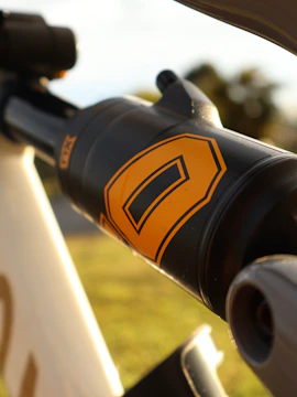 Close-up of a suspension component from a bicycle with a sleek design. The focus is on the black cylinder marked with an orange letter 'E', suggesting a brand or model. The background is blurred, showcasing what seems to be sunlight filtering through trees.