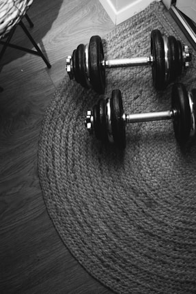 Two adjustable dumbbells are resting on a woven rug placed on a wooden floor. The lighting casts subtle shadows, enhancing the textures of the rug and the wooden floorboards.