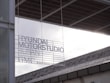 A modern architectural setting features signage with text that reads 'HYUNDAI MOTORSTUDIO BUSAN GMC'. The signage is placed against a backdrop of a contemporary building with visible structural and glass elements. The lighting creates a contrast between the dark edges of the structure and the bright, overcast sky.