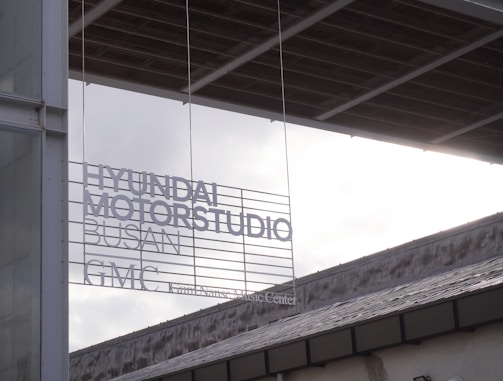 A modern architectural setting features signage with text that reads 'HYUNDAI MOTORSTUDIO BUSAN GMC'. The signage is placed against a backdrop of a contemporary building with visible structural and glass elements. The lighting creates a contrast between the dark edges of the structure and the bright, overcast sky.