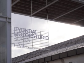 A modern architectural setting features signage with text that reads 'HYUNDAI MOTORSTUDIO BUSAN GMC'. The signage is placed against a backdrop of a contemporary building with visible structural and glass elements. The lighting creates a contrast between the dark edges of the structure and the bright, overcast sky.