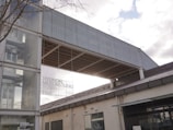 A modern architectural building with a combination of glass and metal elements, featuring a large glass facade with visible text displayed indicating 'Hyundai Motorstudio Busan'. The structure includes a prominent metal screen extending over the side of the building. The sky is partly cloudy.