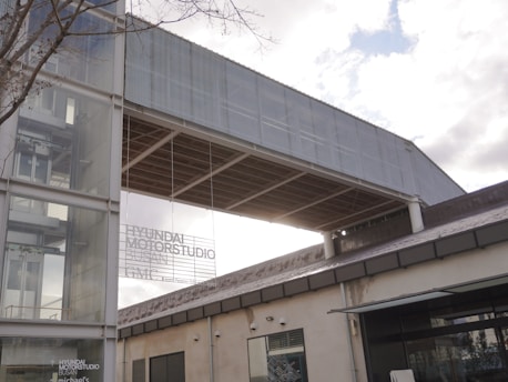 A modern architectural building with a combination of glass and metal elements, featuring a large glass facade with visible text displayed indicating 'Hyundai Motorstudio Busan'. The structure includes a prominent metal screen extending over the side of the building. The sky is partly cloudy.