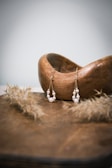 Two pearl earrings hang elegantly from a wooden bowl, with delicate strands of material placed nearby, suggesting a natural and rustic theme.
