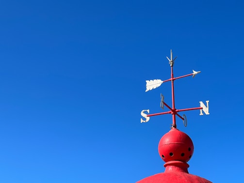 A red weather vane is mounted on a round, red structure against a clear blue sky. The weather vane features directional letters S, N, E, and W.