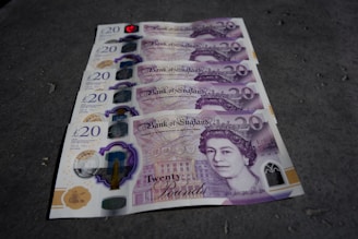 Several £20 British pound banknotes are neatly arranged in a row on a textured surface. The banknotes feature an image of a historical figure and various security features.