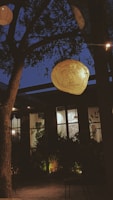 A warmly lit outdoor setting at twilight with a large illuminated sign hanging from a tree branch. The sign is intricately designed, adding an artistic touch. Behind the sign, a building with large windows is visible, partially obscured by leafy plants. The overall scene conveys a cozy and inviting atmosphere.