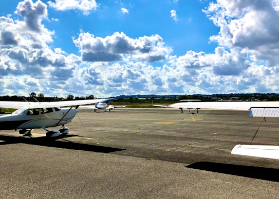 Several light aircraft are parked on a tarmac under a bright, partly cloudy sky. The scene suggests an airfield or small airport with open grassy areas in the distance.