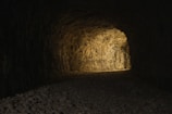 A dimly lit underground tunnel with rough, rocky walls. The tunnel floor is covered with small, irregular stones, leading into darkness.