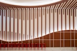 A modern interior features a wall with vertical wooden slats and a wave-like pattern at the top. Below, a row of red stools provides seating, complemented by the white background and polished marble floor.