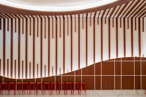 A modern interior features a wall with vertical wooden slats and a wave-like pattern at the top. Below, a row of red stools provides seating, complemented by the white background and polished marble floor.