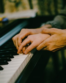 Hands of an adult gently guiding the hands of a child on a piano keyboard, conveying a scene of teaching or assistance.