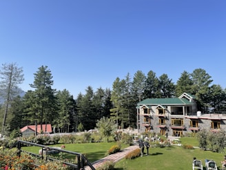 A large building with multiple windows and a roof with a green hue is surrounded by a lush garden. The garden is well-maintained with vibrant flowers and a green lawn. People are seen walking along a pathway bordered by colorful foliage. Tall pine trees are in the background under a clear blue sky.
