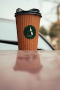 A brown disposable coffee cup with a black lid, featuring a green logo, sits on a reflective surface. The background is slightly blurred, hinting at an outdoor setting with soft, muted colors.