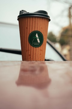 A brown disposable coffee cup with a black lid, featuring a green logo, sits on a reflective surface. The background is slightly blurred, hinting at an outdoor setting with soft, muted colors.