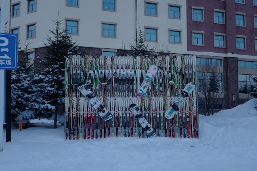 A collection of ski poles and snowboards are arranged vertically against a backdrop, surrounded by snow. The scene is outdoors with evergreen trees and a multi-story building in the background. The snow-covered ground suggests a winter setting. A blue sign with Chinese characters and a parking symbol is visible on the left side.
