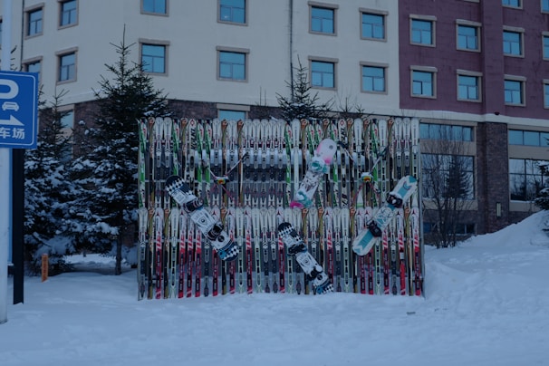 A collection of ski poles and snowboards are arranged vertically against a backdrop, surrounded by snow. The scene is outdoors with evergreen trees and a multi-story building in the background. The snow-covered ground suggests a winter setting. A blue sign with Chinese characters and a parking symbol is visible on the left side.