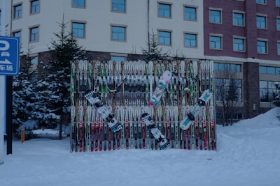 A collection of ski poles and snowboards are arranged vertically against a backdrop, surrounded by snow. The scene is outdoors with evergreen trees and a multi-story building in the background. The snow-covered ground suggests a winter setting. A blue sign with Chinese characters and a parking symbol is visible on the left side.