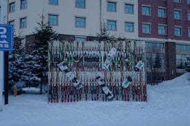 A collection of ski poles and snowboards are arranged vertically against a backdrop, surrounded by snow. The scene is outdoors with evergreen trees and a multi-story building in the background. The snow-covered ground suggests a winter setting. A blue sign with Chinese characters and a parking symbol is visible on the left side.