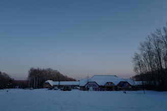 A winter landscape featuring a snow-covered ground in front of a building with a few cars and a truck parked nearby. The building has a large roof and there are several trees with bare branches surrounding the area, set against a clear evening sky.