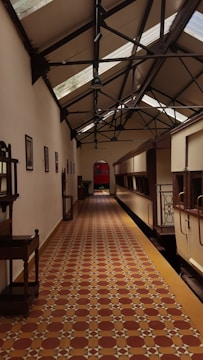 A long corridor with patterned tile flooring and a high ceiling supported by metal beams. The walls are adorned with framed pictures, and there are several pieces of furniture along the left side. A series of skylights allow natural light to illuminate the space. At the far end, a door or window features a colorful, translucent panel.
