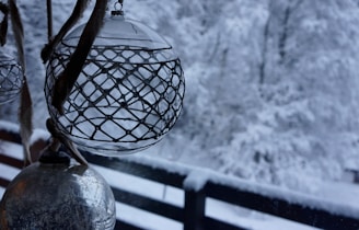 Ornate glass ornaments hang indoors near a window, with a snowy winter landscape visible outside. The intricate design on the ornaments contrasts with the soft, blurred background of snow-covered trees and a wooden fence.