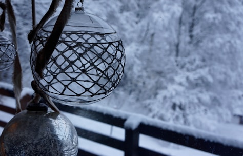 Ornate glass ornaments hang indoors near a window, with a snowy winter landscape visible outside. The intricate design on the ornaments contrasts with the soft, blurred background of snow-covered trees and a wooden fence.