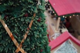 A large, decorated Christmas tree with green pine branches and red berries is prominently featured in the foreground. The tree is adorned with metallic gold ribbons. In the background, there are red-roofed stalls, resembling a holiday market setting.