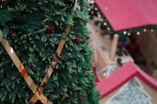 A large, decorated Christmas tree with green pine branches and red berries is prominently featured in the foreground. The tree is adorned with metallic gold ribbons. In the background, there are red-roofed stalls, resembling a holiday market setting.