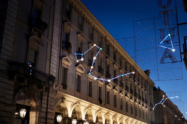 A cityscape at dusk features a row of classic European-style buildings adorned with ornate architectural details and balconies. Illuminated constellations are suspended above the street, adding a whimsical touch under the deepening blue sky. Street lamps cast a warm glow on the scene, contributing to a serene and elegant atmosphere.