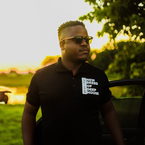 A person wearing sunglasses is standing outdoors during sunset, with a look of confidence. They are dressed in a black polo shirt with the text 'NEW BREED OF DEEP HOUSE' printed on it. Behind them, a car door is slightly open, and there are lush green trees and grass illuminated by the golden sunlight.