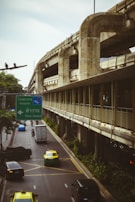 A busy urban road is flanked by an elevated concrete structure, possibly a metro or railway track. Several vehicles, including cars, taxis, and motorbikes, navigate the street. A prominent green road sign provides directions to a hospital and police station in both Thai and English. The scene is surrounded by greenery, and the atmosphere feels overcast.